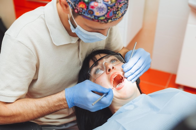 남자 의사는 건강한 치아의 개념 옆에 있는 사무실에서 치과 의사의 의자에 앉아 있는 젊은 환자의 구강을 검사합니다
