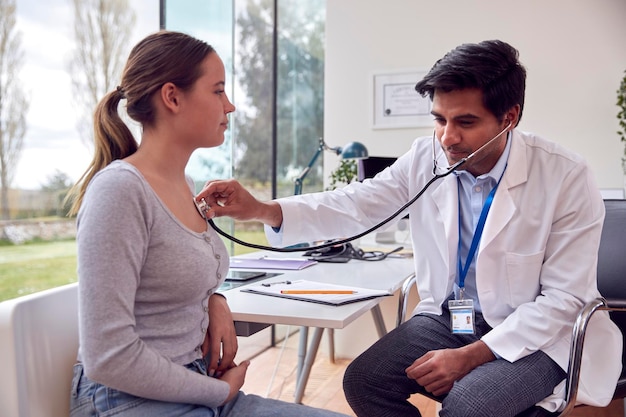 Male Doctor Or GP Wearing White Coat Examining Teenage Girl Listening To Chest With Stethoscope