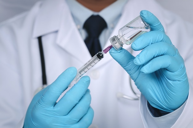 Male doctor hands on blue gloves with stethoscope on shoulder holding syringe and Covid-19 coronavirus vaccine vial or drug on white wall. Healthcare And Medical concept