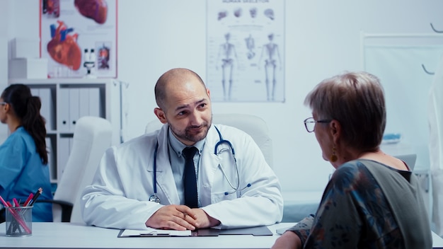 Male doctor interviewing an elderly woman in his cabinet. Medical staff working in modern private modern healthcare system, visit specialist appointment. Treatment medical prescription from therapist