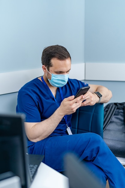 携帯電話を見ているオフィスのマスクの男性医師。現代の病院のオフィスの背景。