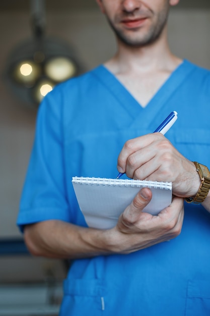 Врач-мужчина в медицинской синей стерильной одежде стоит в палате больницы и пишет в блокноте