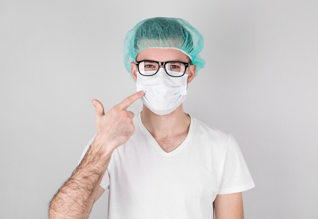 Male doctor in medical uniform points his finger at a medical mask on his face.