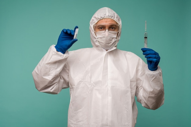 A male doctor in protective overalls and a mask holds an ampoule with a vaccine and syringe in his hands