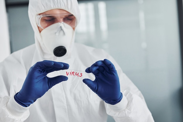 male doctor scientist in lab coat, defensive eyewear and mask holds glass with virus word on it