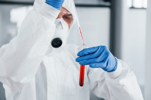 Male doctor scientist in lab coat, defensive eyewear and mask works with test tube with blood inside of it