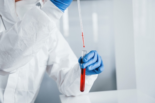 Male doctor scientist in lab coat, defensive eyewear and mask works with test tube with blood inside of it