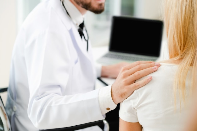 Male doctor talks to female patient in hospital office while looking at data on laptop computer.