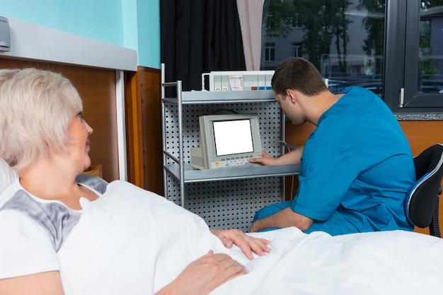 Male doctor in uniform is looking at monitor of medical device while female patient is lying in the hospital bed in the hospital ward. Healthcare concept