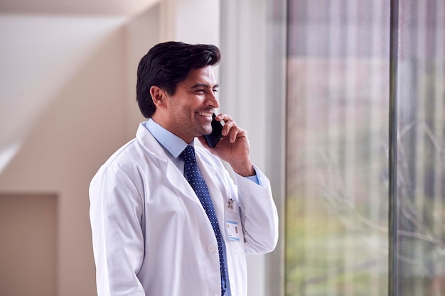 Мужчина-врач в белом халате стоит в больничном коридоре и разговаривает по мобильному телефону