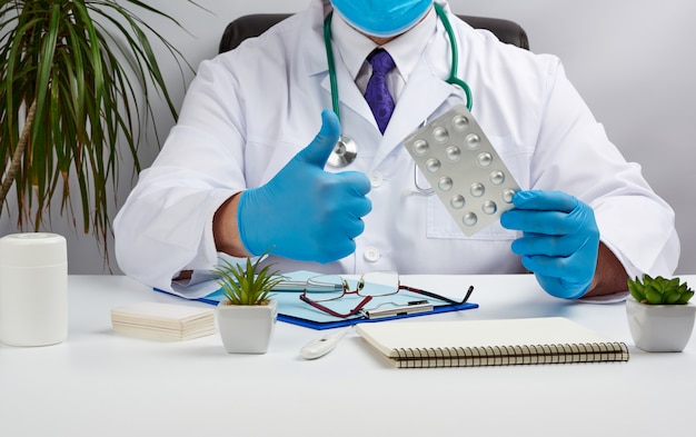 Male doctor in a white coat and blue latex gloves sits at a white desk in his office and shows a pack of pills in a blister