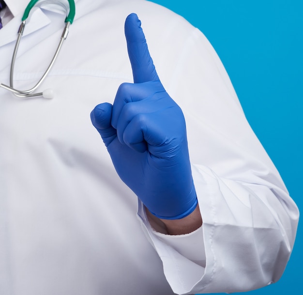 Male doctor in white coat, blue medical gloves shows hand gesture of attention, index finger up