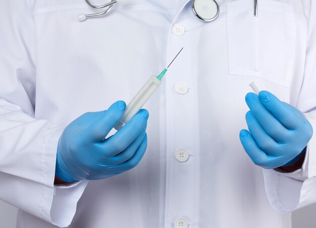 Male doctor in a white coat and tie stands and holds a plastic syringe with a needle on a white background, wearing blue sterile medical gloves