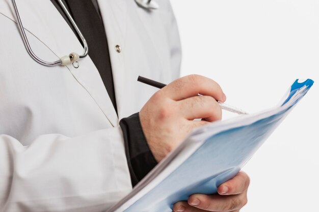 A male doctor in a white coat with a stethoscope fills out a medical history. White wall.