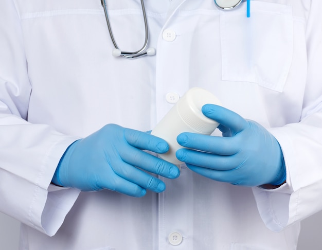 Male doctor in a white medical coat and blue latex gloves holds a white plastic jar of pills