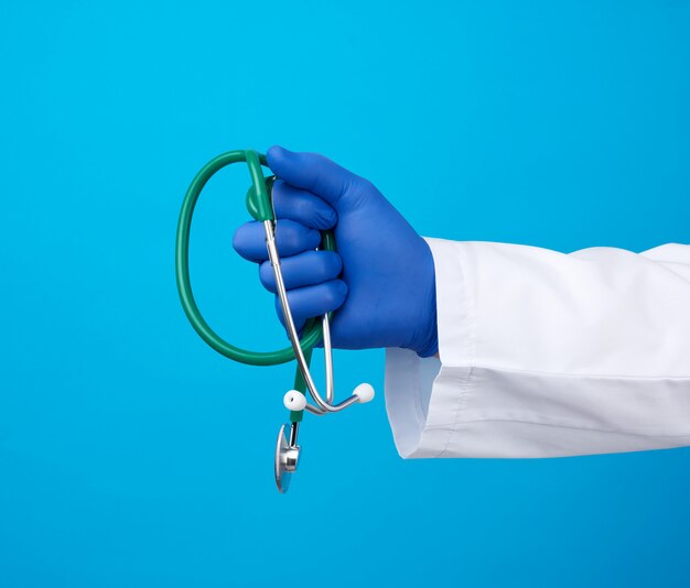 Male doctor in a white medical coat stands and holds a green stethoscope on a blue background