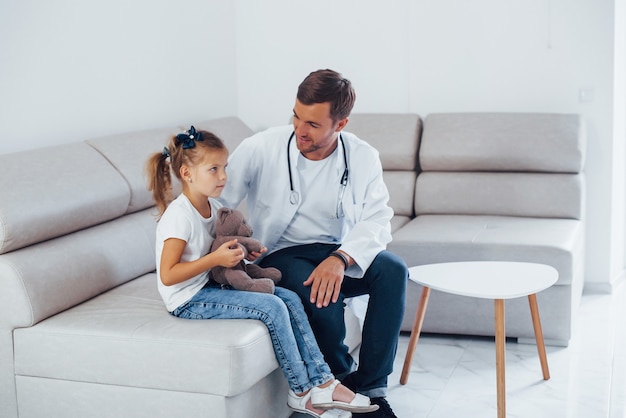 Male doctor in white uniform sits in the clinic with little girl.