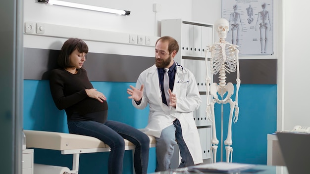 Male doctor and woman with pregnancy belly doing medical consultation at checkup appointment. Physician examining pregnant patient, giving advice about childbirth and health care.