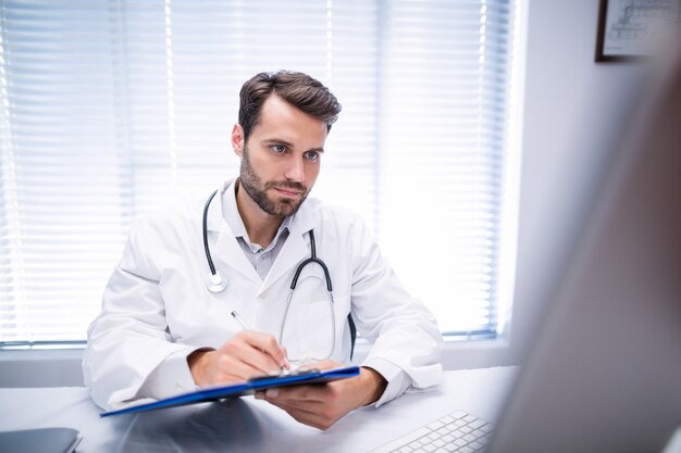 Male doctor writing on clipboard while working on personal computer