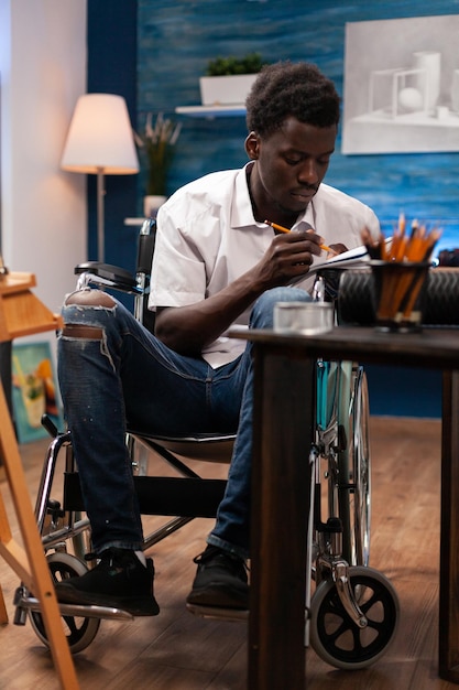 Male drawing professional wheelchair user writing notes with\
paper and pencil on desk in bedroom. african american artist living\
with medical condition making hand drawn sketch with colored\
pencil.