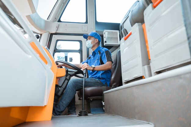 A male driver in uniform and wearing a mask while holding the steering wheel and gear lever in the bus