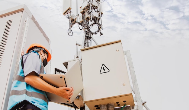 Lavoratore elettronico maschio che utilizza il computer portatile per ispezionare la torre del segnale del sistema del circuito di telecomunicazione.