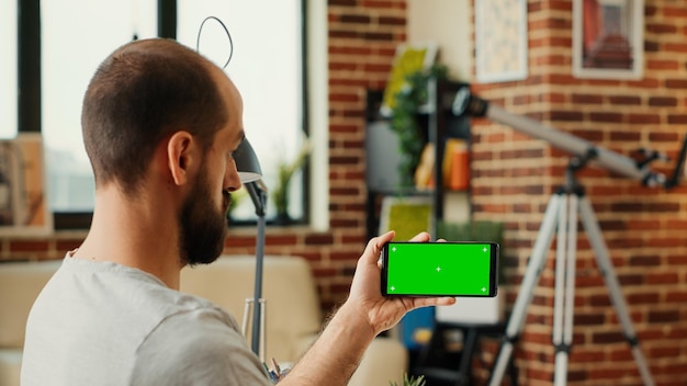 Male employee checking horizontal greenscreen display on mobile phone, looking at smartphone with isolated chroma key template. Working with blank mockup copy space background.