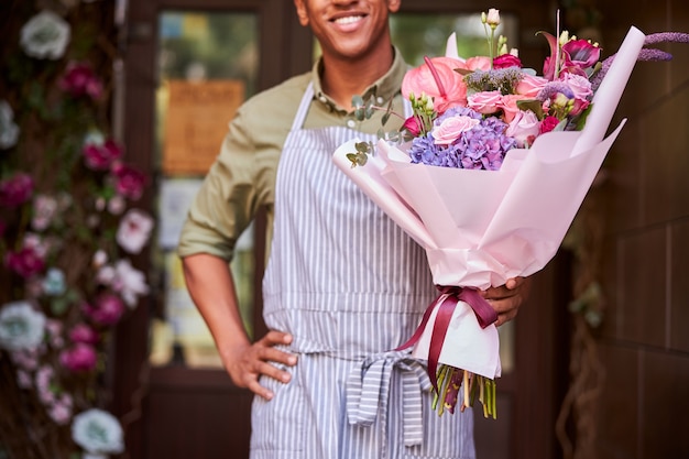 Male employee standing at the store threshold with a bunch of flowers keeping his hand on a hip