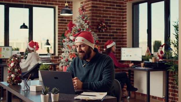 Foto impiegato maschio che parla in videoconferenza online, indossando il cappello di babbo natale in ufficio festivo. partecipare a una riunione di teleconferenza remota durante le vacanze natalizie, conferenza remota.