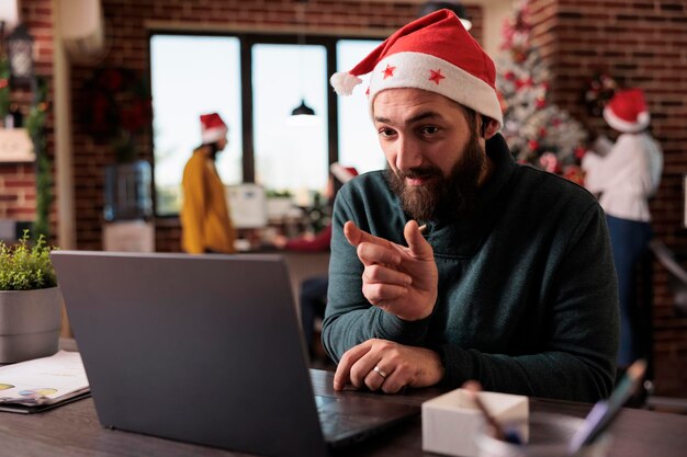 Male employee talking on videoconference call meeting in business office with festive decorations and christmas tree. Using remote online teleconference to chat during holiday season.