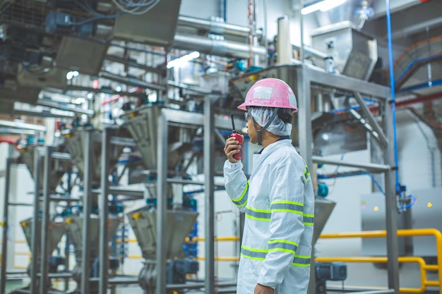 Photo male engineers sound protection and uniform staffhand holding portable radio transceiver for communication work inspection process powder cellar at the with vertical stainless steel tanks factory