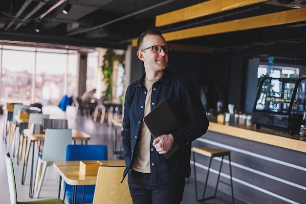 Photo male entrepreneur in black shirt and glasses standing in office with laptop in hand successful corporate boss feeling good from rich lifestyle