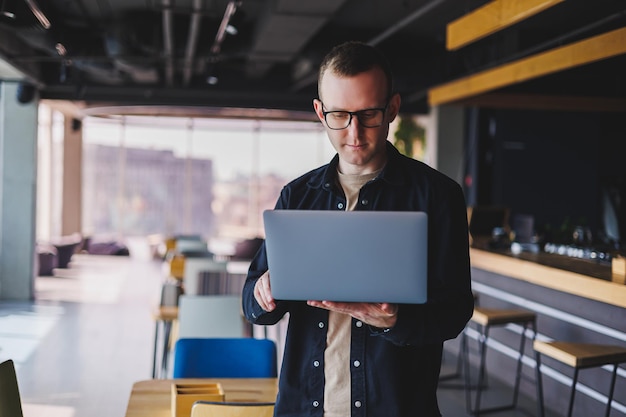 Male entrepreneur in black shirt and glasses standing in office with laptop in hand successful corporate boss feeling good from rich lifestyle