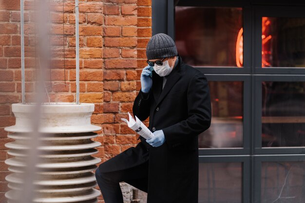 L'imprenditore maschio discute degli emittenti di lavoro tramite smartphone, indossa maschera e guanti protettivi, tiene documenti, pone il muro di mattoni. virus epidemico. nuovo concetto di coronavirus. rimani sano e salvo