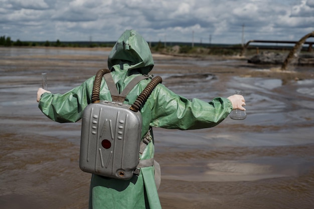 A male environmentalist in a green protective suit and gas mask takes a sample of water in a polluted lake Waste from production A scientist does a toxicological study of toxic waste