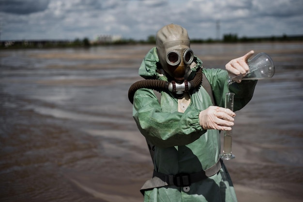 A male environmentalist in a green protective suit and gas mask takes a sample of water in a polluted lake Waste from production A scientist does a toxicological study of toxic waste