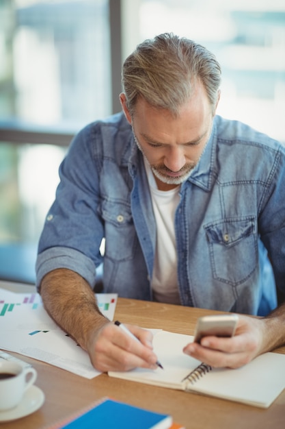 Male executive writing in organizer while using mobile phone