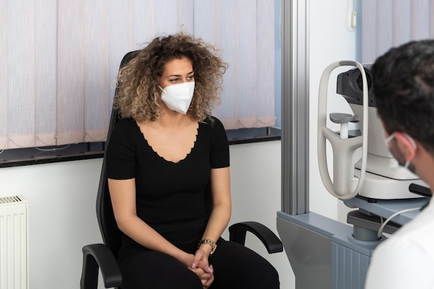 Male Eye Doctor Listen Carefully Young Woman Wearing Surgical Medical Mask to Prevent Infection in Office Portrait