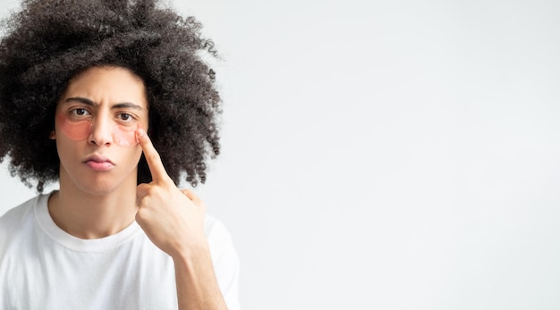 Male facial care Antiaging procedure Metrosexual lifestyle Unhappy tired handsome guy with brown curly hair pointing at pink hydrogel eye patches on face isolated on light copy space background