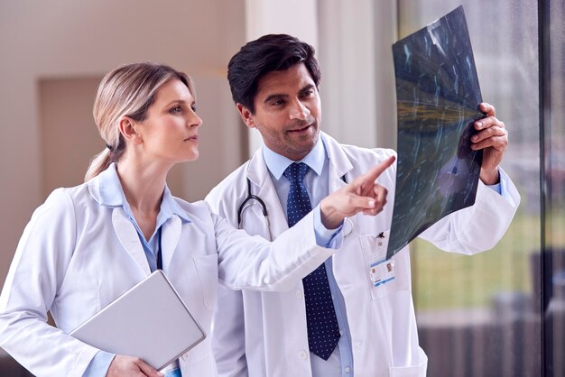 Male And Female Doctors Wearing White Coats Standing In Hospital Corridor Looking At CT Or MRI Scan