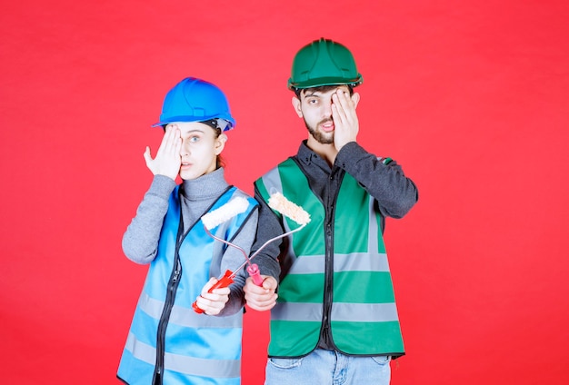 Male and female engineers with helmet holding trim rollers and look thrilled and stressed.