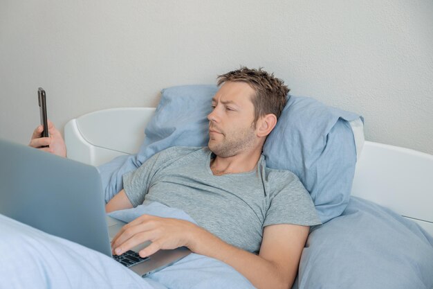 Male freelancer checking email on smartphone while working with laptop in bed in the morning man