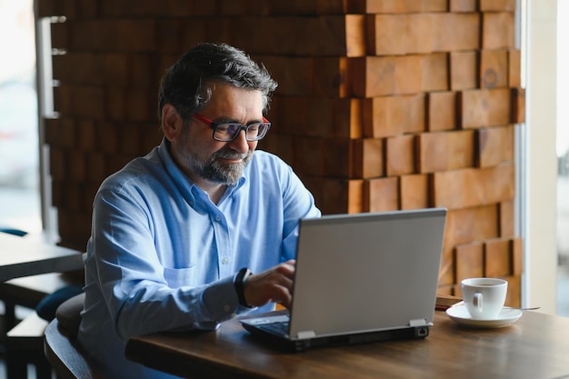 Male freelancer is working in a cafe on a new business project Sits at a large window at the table Looks at a laptop screen with a cup of coffee