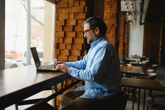 남성 프리랜서는 새로운 비즈니스 프로젝트를 위해 카페에서 일하고 있습니다. 테이블의 큰 창에 앉아 커피 한 잔과 함께 노트북 화면을 봅니다.