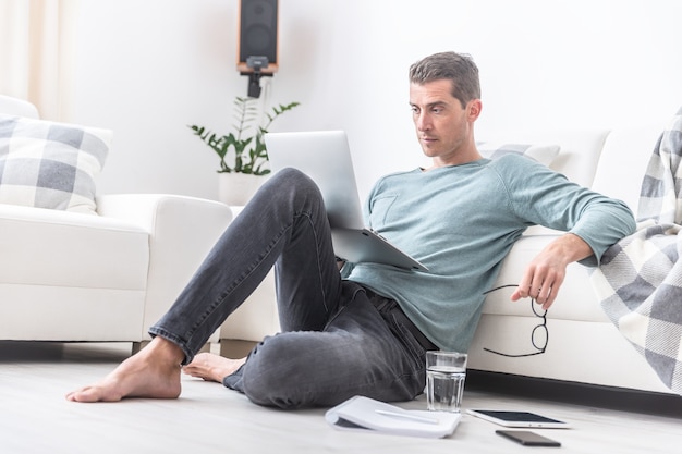 Male freelancer sits barefoot on the living room floor working on a laptop.