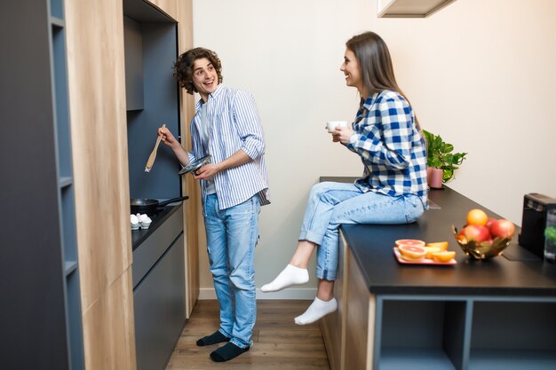 Male fry in a frying pan and his woman sitting on the table and drinking coffee