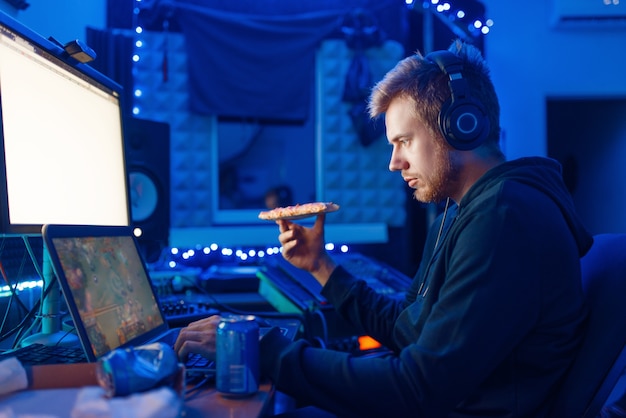 Male gamer drinking an energy drink at his workplace with laptop and desktop PC, gaming night lifestyle. Computer games player in his room with neon light, streamer