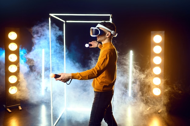Фото Геймер-мужчина играет в игру, используя гарнитуру виртуальной реальности и геймпад в светящемся кубе. темный интерьер игрового клуба, технология vr с 3d-зрением