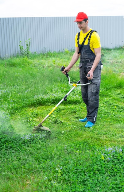 男性の庭師が裏庭の芝生の緑の草を刈り取り、庭の手入れのためにガソリン芝刈り機トリマーを使用