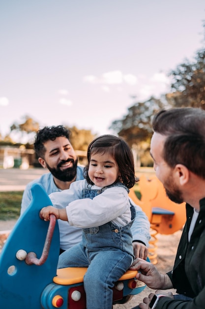 Male gay couple playing with their daughter on a seesaw in the park Modern family concept Vertical